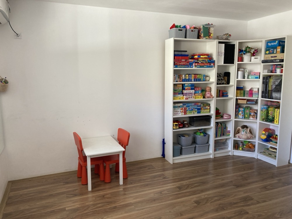 Sala de terapie: măsuță și scăunele portocalii pentru copii, lângă rafturile cu jocuri